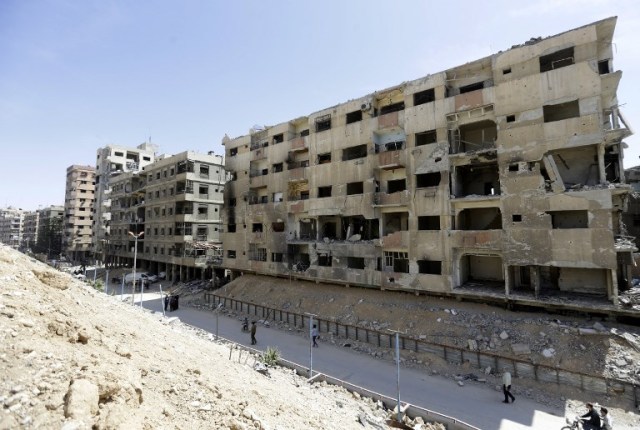 Los sirios caminan frente a edificios dañados en Douma en las afueras de Damasco el 16 de abril de 2018 durante una gira organizada después de que el ejército sirio declarara que todas las fuerzas contrarias al régimen habían abandonado Eastern Ghouta, tras una ofensiva de dos meses en el enclave rebelde. El anuncio, que representa una victoria estratégica clave para el presidente Bashar al-Assad, se produjo horas después de que los ataques liderados por Estados Unidos golpearan objetivos del gobierno sirio en respuesta a un presunto ataque químico en Douma, la principal ciudad del enclave. / AFP PHOTO / LOUAI BESHARA